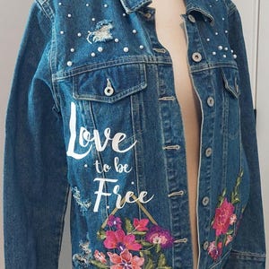 May include: A blue denim jacket with pearl accents and floral embroidery. The jacket features the text "Love to be Free" in white script. The jacket has a distressed look with a torn section on the upper left side.