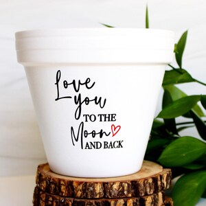 May include: A white ceramic flower pot with the text "Love you to the moon and back" in black script. A small red heart accents the text. The pot sits on a rustic wooden base, with green foliage in the background.