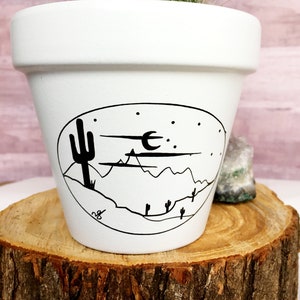 May include: White ceramic planter with a black and white illustration of a desert landscape with a cactus, mountains, a crescent moon, and stars. A small cactus with pink flowers is planted in the pot.