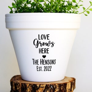 May include: White ceramic planter with the text "Love Grows Here" and a small heart, followed by "The Hensons Est. 2022". A green plant is growing in the planter, which is sitting on a stack of wooden rounds.
