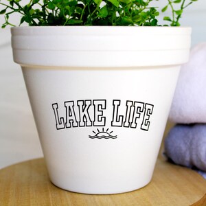 May include: White ceramic planter with the text "LAKE LIFE" and a small sun graphic. The planter is filled with green leafy plants.