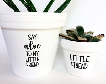 May include: Two white ceramic plant pots with black text. The larger pot has an aloe vera plant, and the text reads "SAY aloe TO MY LITTLE FRIEND." The smaller pot has a succulent and reads "LITTLE FRIEND."
