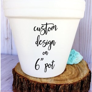 May include: White ceramic flower pot with the text "custom design on 6" pot" written in black. The pot is sitting on a wooden slice.