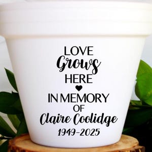 May include: White ceramic flower pot with the text "LOVE GROWS HERE IN MEMORY OF Claire Coolidge 1949-2025". The pot is sitting on a wooden slice with green leaves in the background.