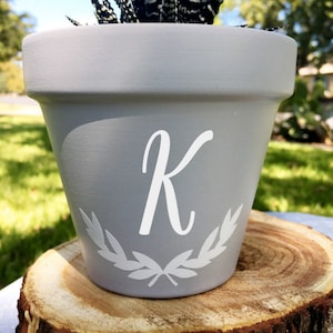 May include: A gray terracotta planter with a white letter K and a white wreath design. The planter is filled with a green succulent plant.