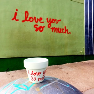 May include: A white ceramic planter with the words "i love you so much" written in red paint. The planter is sitting on a gray, textured surface.