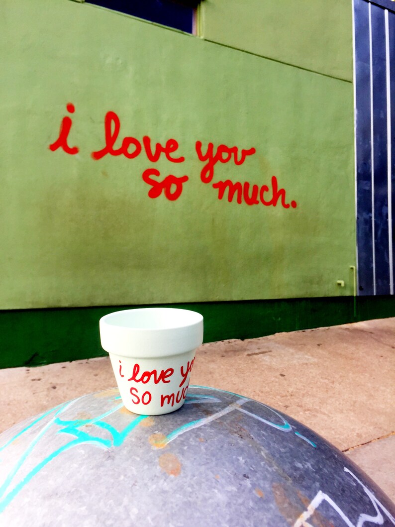 May include: A white ceramic planter with the words "i love you so much" written in red paint. The planter is sitting on a gray, textured surface.