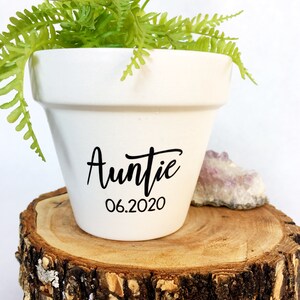 May include: White ceramic planter with the word "Auntie" and the date "06.2020" written in black. A green fern plant is growing in the planter, which is sitting on a wooden log slice.