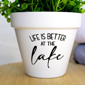 May include: A white ceramic flower pot with the text "LIFE IS BETTER AT THE lake" in black script. The pot is filled with green leafy plants and sits on a wooden surface.
