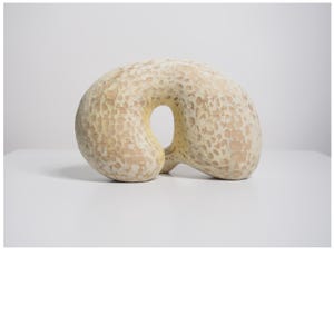 May include: A cream-colored, abstract sculpture with a large central opening. The textured surface has a pattern of small, irregular indentations. The form is organic and rounded, suggesting a natural or handcrafted origin. The sculpture sits on a white surface.
