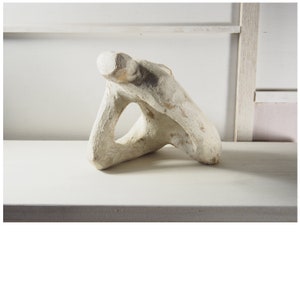 Op de afbeelding: Witte keramische sculptuur van een abstract menselijk figuur met een gat in het midden. De sculptuur staat op een wit oppervlak.