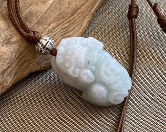 Puede incluir: Un colgante de jade blanco tallado en forma de criatura mítica, con detalles intrincados. El colgante está unido a un cordón marrón con una cuenta de plata.