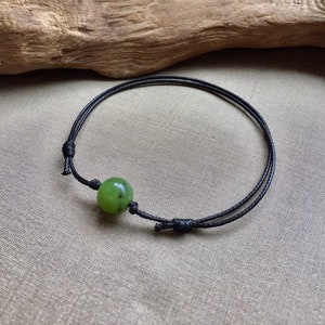 May include: A black cord bracelet featuring a round, green jade bead. The bracelet is knotted for adjustability. The background is a neutral tan color, with a piece of driftwood in the upper left corner.
