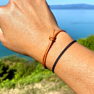 May include: Two bracelets on a wrist. One is a brown leather bracelet with a knot closure. The other is a black elastic bracelet.