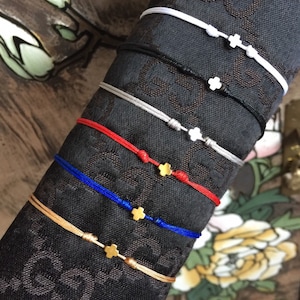 May include: A set of six bracelets with different colored cords and gold cross charms. The bracelets are arranged in a stack, with the white cord bracelet on top and the gold cord bracelet on the bottom.
