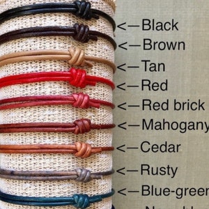 May include: A display of ten leather cord bracelets in various colors, including black, brown, tan, red, red brick, mahogany, cedar, rusty, blue-green, and navy. The bracelets are arranged vertically on a fabric background.
