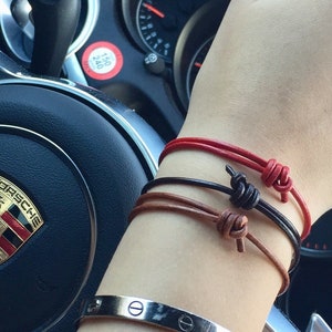 May include: A person's wrist with three leather bracelets in red, brown, and black, tied with knots. A silver bracelet with a screw design is also on the wrist.