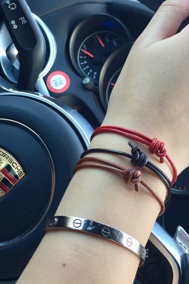 May include: A person's wrist with three leather bracelets in red, brown, and black, tied with knots. A silver bracelet with a screw design is also on the wrist.