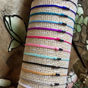 Puede incluir: Una colección de pulseras de cuerda de colores con pequeñas cuentas negras. Las pulseras están dispuestas en una pila sobre un fondo de tela marrón.