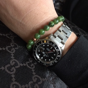 May include: A wrist featuring a silver wristwatch with a black face and a green beaded bracelet. The watch has a black bezel with white numerals. The bracelet has green beads with black speckles.