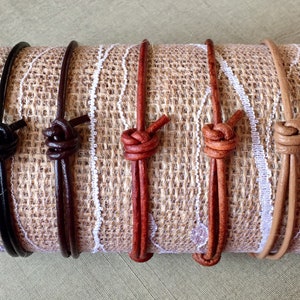 May include: Five leather bracelets in different colors, including black, brown, red, tan, and natural. The bracelets are tied with a knot and are displayed on a burlap background.