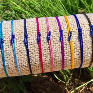 May include: A collection of 14 colorful bracelets with blue beads. The bracelets are made of thin cord and are displayed on a woven surface.
