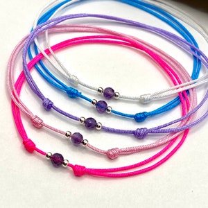 Könnte beinhalten: Fünf bunte Armbänder aus Kordel mit Amethyst-Perlen und silbernen Akzenten. Die Armbänder sind auf einer weißen Oberfläche übereinander gelegt.