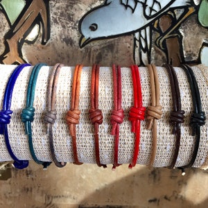 May include: Ten leather bracelets in various colors, including blue, green, brown, orange, red, and black. The bracelets are tied with a knot and are displayed on a white fabric background.