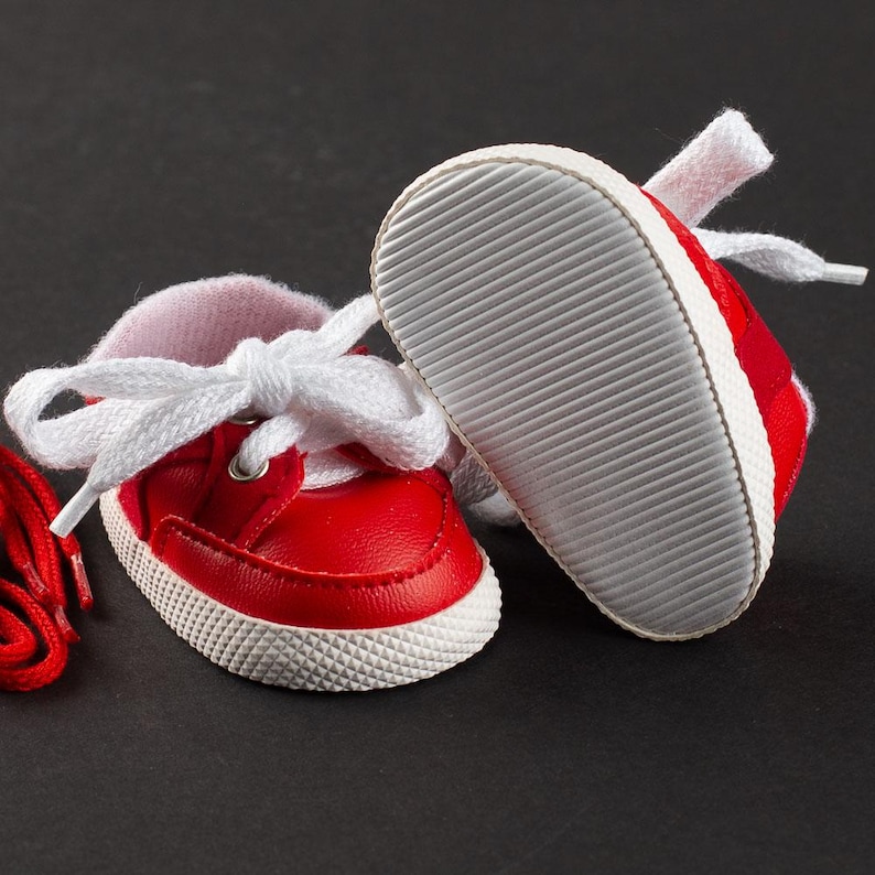 Op de afbeelding: Een paar kleine rode babysneakers met witte veters en zolen. De schoenen hebben een klassiek ontwerp, met een rood bovenwerk, witte rand en een getextureerde witte zool. De veters zijn gestrikt.