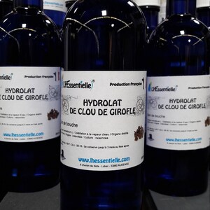 Puede incluir: Primer plano de tres botellas de vidrio azul con tapas blancas. Las botellas est&aacute;n etiquetadas con "Hydrolat de Clou de Girofle" y "L'Essentielle". Las botellas est&aacute;n sobre una superficie de madera.