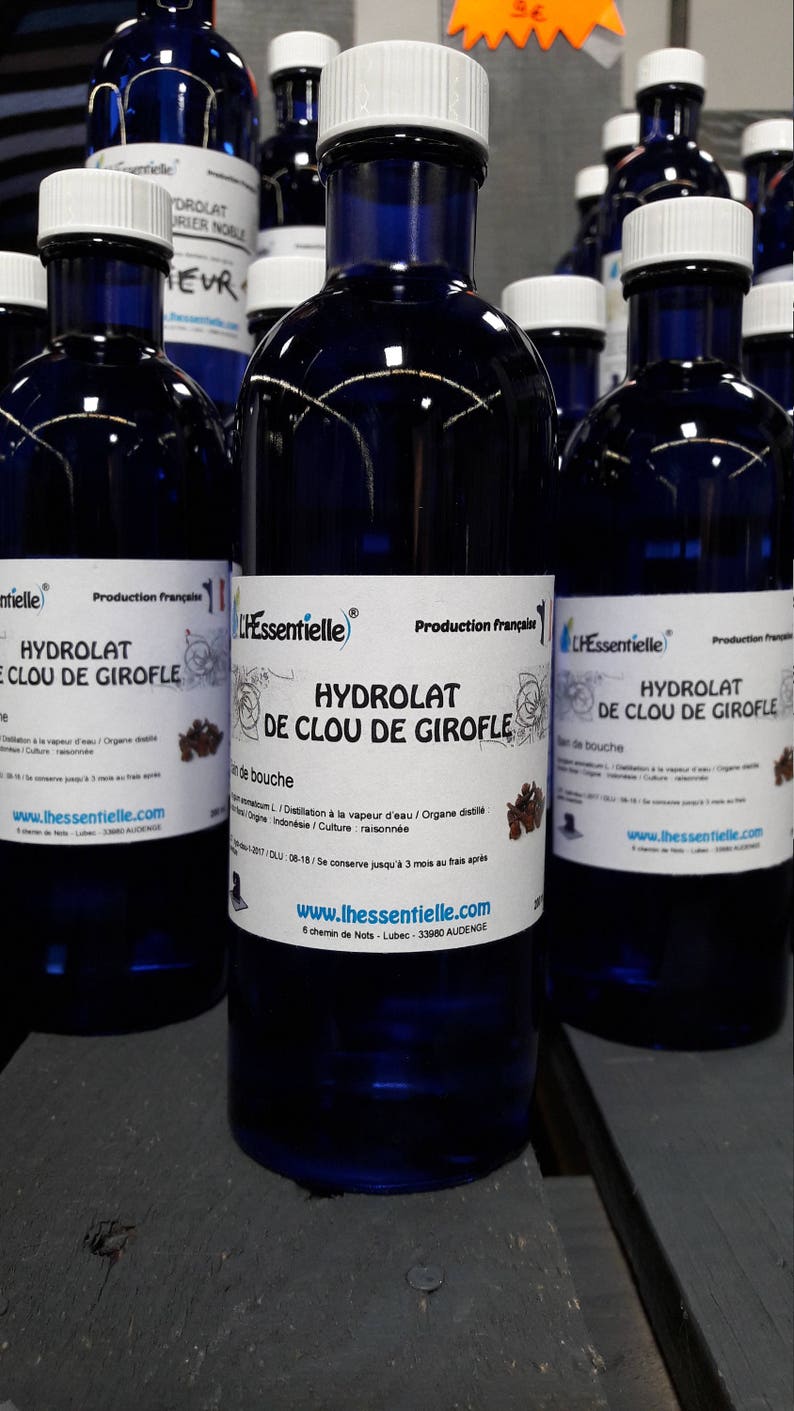 Puede incluir: Primer plano de tres botellas de vidrio azul con tapas blancas. Las botellas est&aacute;n etiquetadas con "Hydrolat de Clou de Girofle" y "L'Essentielle". Las botellas est&aacute;n sobre una superficie de madera.