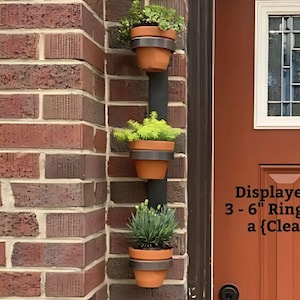 May include: Three black metal plant holders with a clear coat finish, each holding a potted plant. The holders are mounted on a brick wall next to a red door with a glass window. The text "MAIL" is on a black sign below the holders. The text "Displayed in 30" featuring 3-6" Ring Plant Holders with a {Clear Coated Finish)." is below the door.