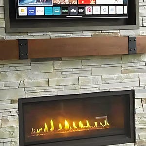 May include: A modern fireplace with a black frame and a realistic flame effect. The fireplace is built into a wall of stacked stone with a wooden mantel above it. A flat screen TV is mounted above the mantel.