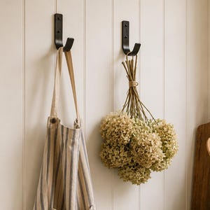 May include: Two black metal wall hooks mounted on a white paneled wall. One hook holds a beige and blue striped apron, while the other holds a bundle of dried hydrangea flowers tied with twine. A wooden bowl with artichokes sits on the counter.
