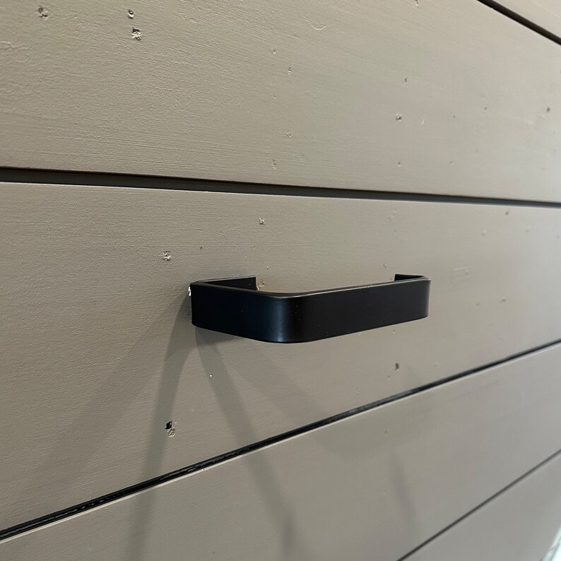 May include: A black metal cabinet door handle with a rectangular shape.