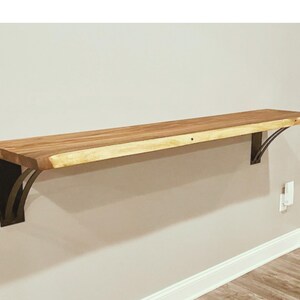 May include: A wooden shelf with live edge detailing is mounted to a wall with two black metal brackets. The shelf is made of natural wood with a light brown finish.