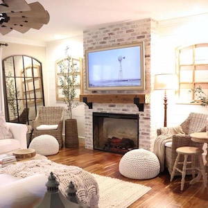 May include: A cozy living room with a brick fireplace, a large framed television, and wicker furniture. The room features a neutral color palette with wooden floors, a woven rug, and decorative poufs, creating a warm and inviting atmosphere.