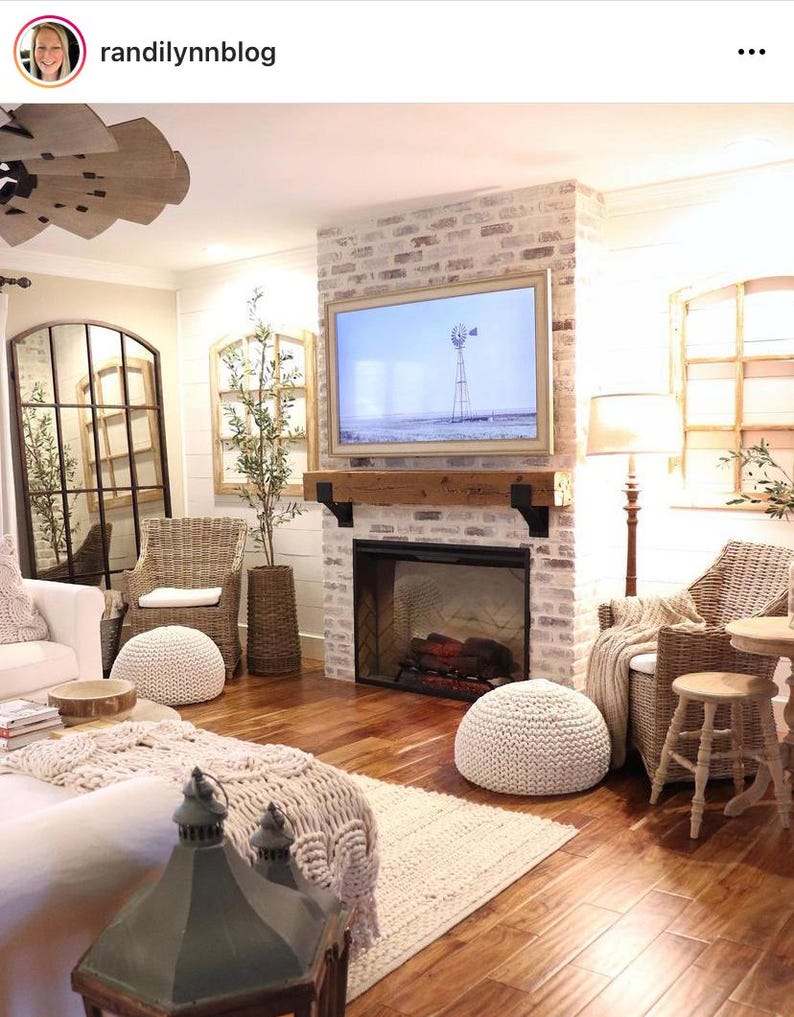 May include: A cozy living room with a brick fireplace, a large framed television, and wicker furniture. The room features a neutral color palette with wooden floors, a woven rug, and decorative poufs, creating a warm and inviting atmosphere.