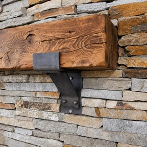 May include: A thick, rectangular wooden beam with a prominent knot and visible wood grain, supported by a dark gray metal bracket. The bracket features a flat top plate, a curved support arm, and a vertical base plate secured with metal bolts.