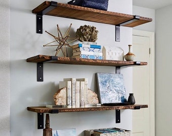 May include: Four wooden shelves with black metal brackets, mounted on a white wall. The shelves are decorated with books, a gold metal sculpture, a coral sculpture, a white ceramic house, a white ceramic vase, a blue and white painting, a wooden sculpture, a silver sculpture, and a white wicker basket.
