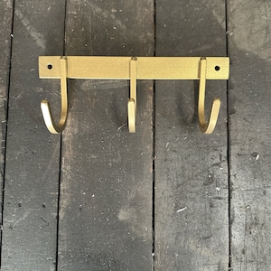 May include: Gold metal wall mounted hook rack with three hooks. The rack is mounted on a dark wood surface.