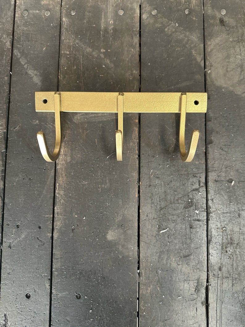 May include: Gold metal wall mounted hook rack with three hooks. The rack is mounted on a dark wood surface.