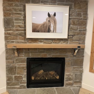 May include: A framed photograph of a horse with long, flowing mane and forelock hangs above a fireplace. The fireplace is constructed of stone and features a wooden mantel and a black firebox with faux logs.