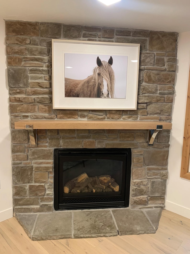 May include: A framed photograph of a horse with long, flowing mane and forelock hangs above a fireplace. The fireplace is constructed of stone and features a wooden mantel and a black firebox with faux logs.