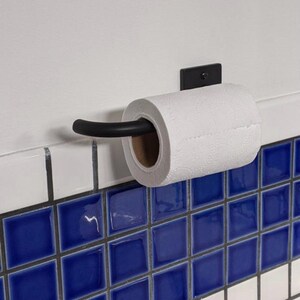May include: Black metal toilet paper holder mounted on a white tiled wall with blue and white square tiles. A roll of white toilet paper is on the holder.