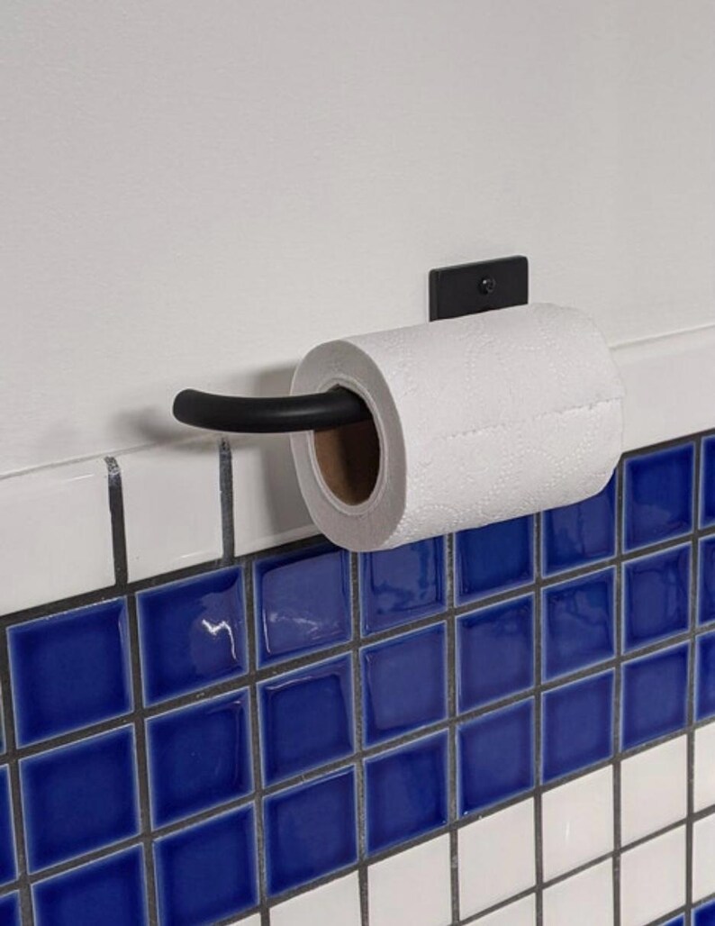 May include: Black metal toilet paper holder mounted on a white tiled wall with blue and white square tiles. A roll of white toilet paper is on the holder.
