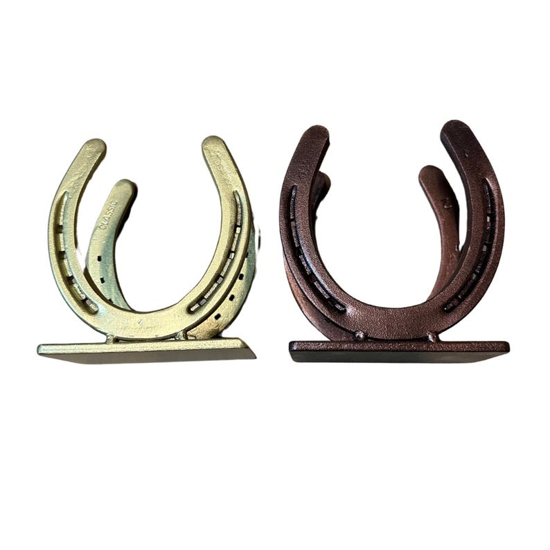 May include: Two U-shaped metal horseshoe holders, one finished in a metallic gold tone and the other in a dark bronze tone. Each piece features a flat rectangular base and a curved frame with visible nail holes, designed to stand upright.