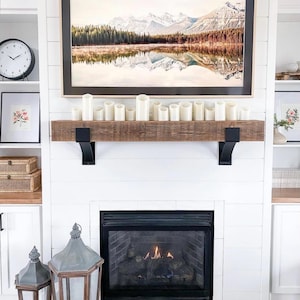 May include: A cozy living room scene featuring a white fireplace with a black firebox. A wooden mantel holds white candles. Above, a framed landscape painting depicts mountains and a lake. Decorative lanterns and a clock complete the decor.