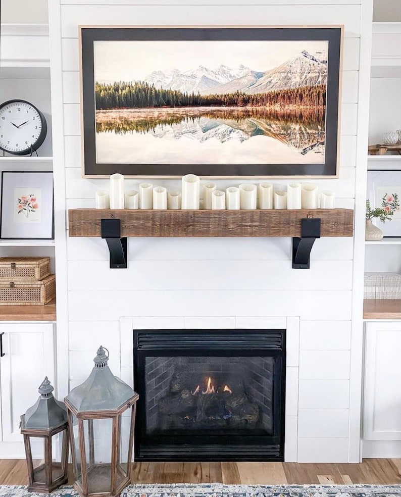 May include: A cozy living room scene featuring a white fireplace with a black firebox. A wooden mantel holds white candles. Above, a framed landscape painting depicts mountains and a lake. Decorative lanterns and a clock complete the decor.