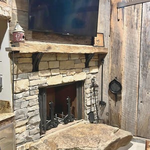 May include: A stone fireplace with a rustic wooden mantel and a black metal fireplace screen. The fireplace is surrounded by a stone wall and has a large flat stone hearth.
